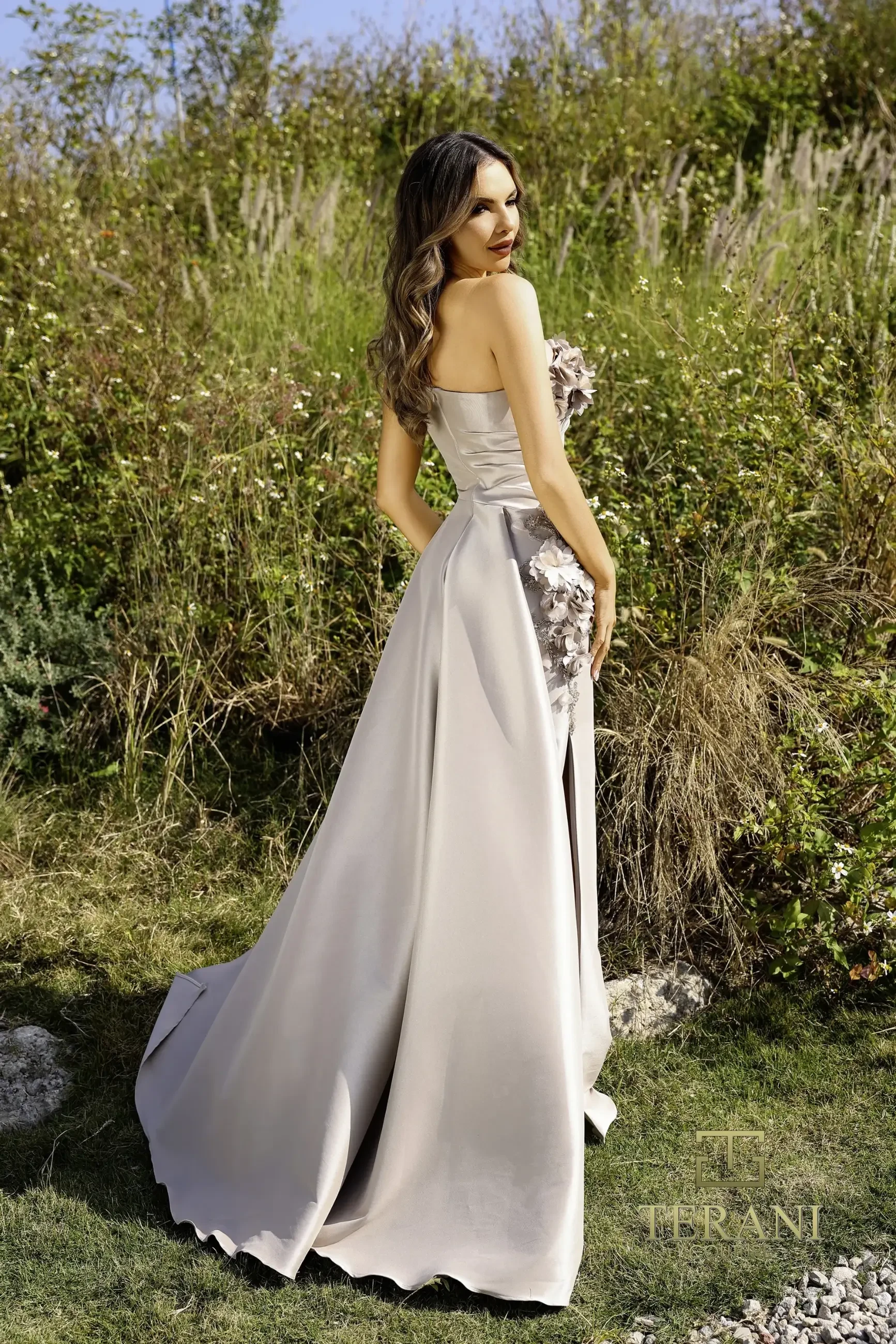 Model is wearing the Terani 252E5116, a taupe mock wrap gown with dramatic draping, metallic jacquard shimmer, delicate bead detailing, and an attached overskirt.
