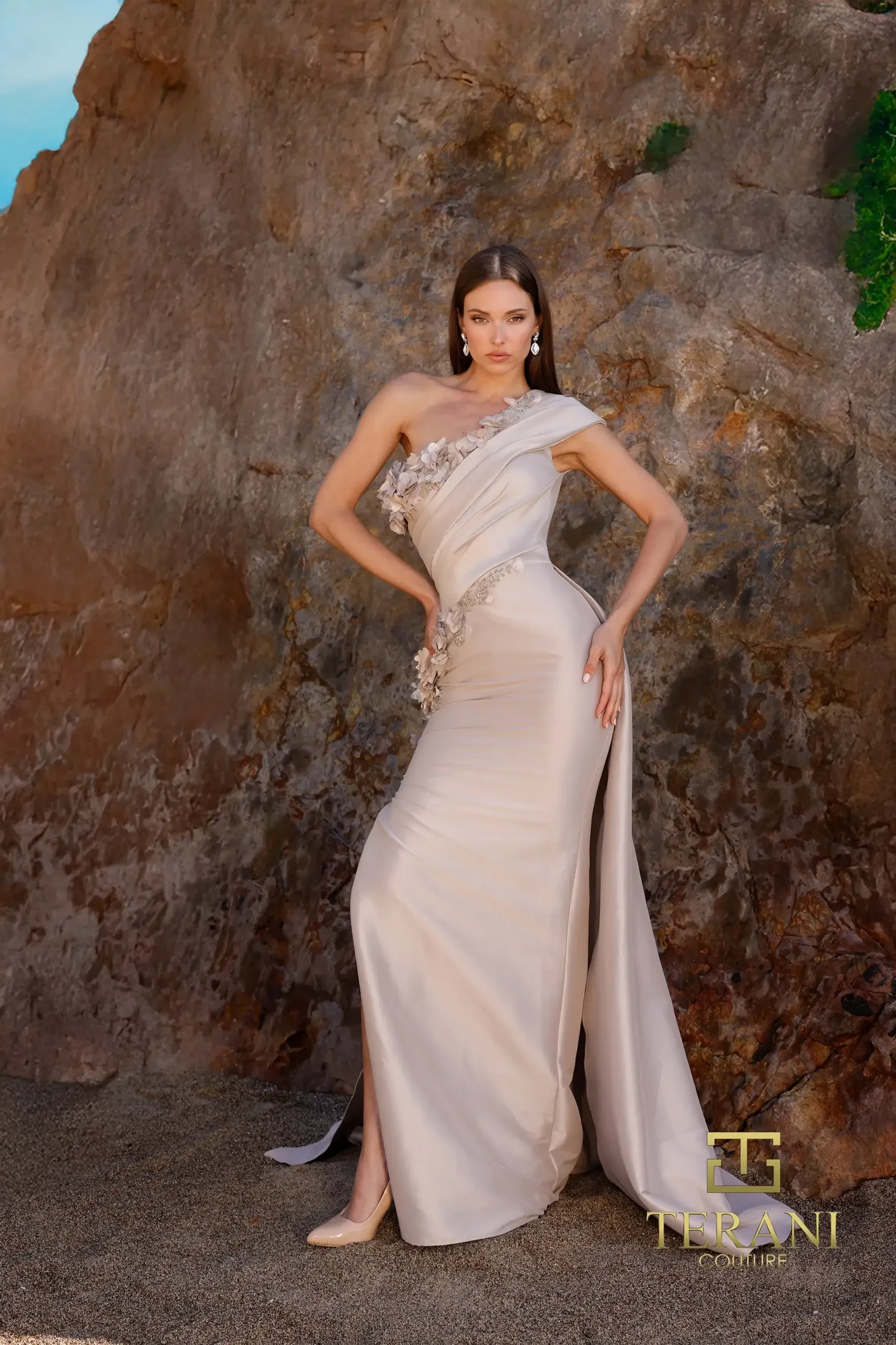 Model is wearing the Terani 252E5116, a taupe mock wrap gown with dramatic draping, metallic jacquard shimmer, delicate bead detailing, and an attached overskirt.