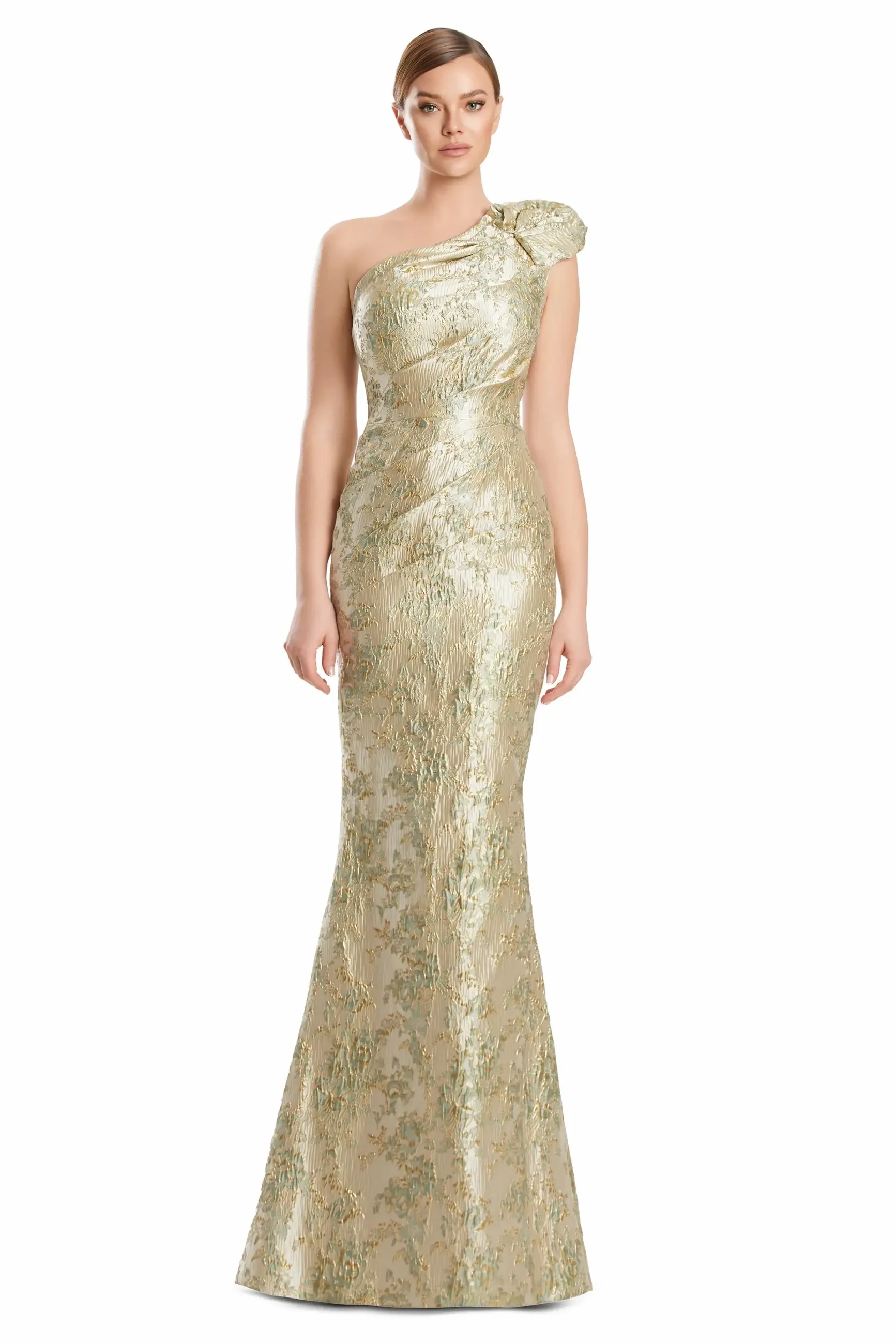 A woman is wearing a formal, one-shoulder gown that features a textured, shimmery light gold fabric and a floral embellishment on the shoulder. The dress has a fitted silhouette that flares towards the bottom.
