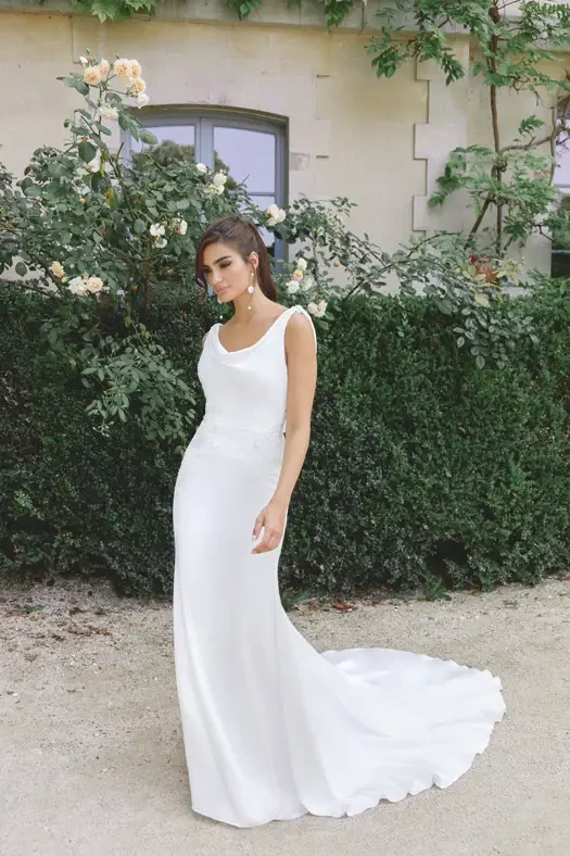 A woman in a white gown stands elegantly in a garden with green shrubs and flowers in the background.