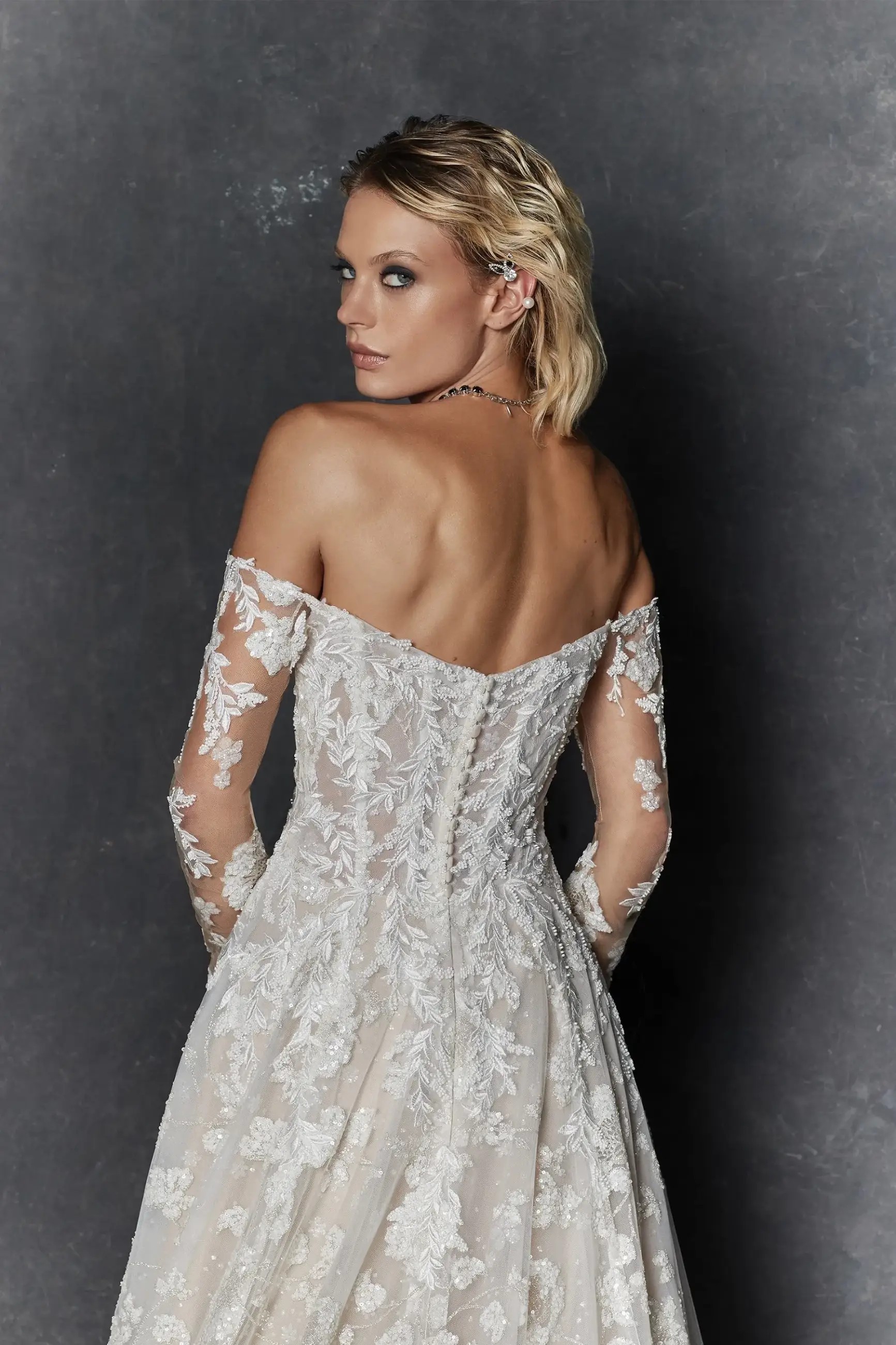 Model is wearing an a-line wedding dress with floral glitter tulle and a plunging off the shoulder neckline and long sheer sleeves