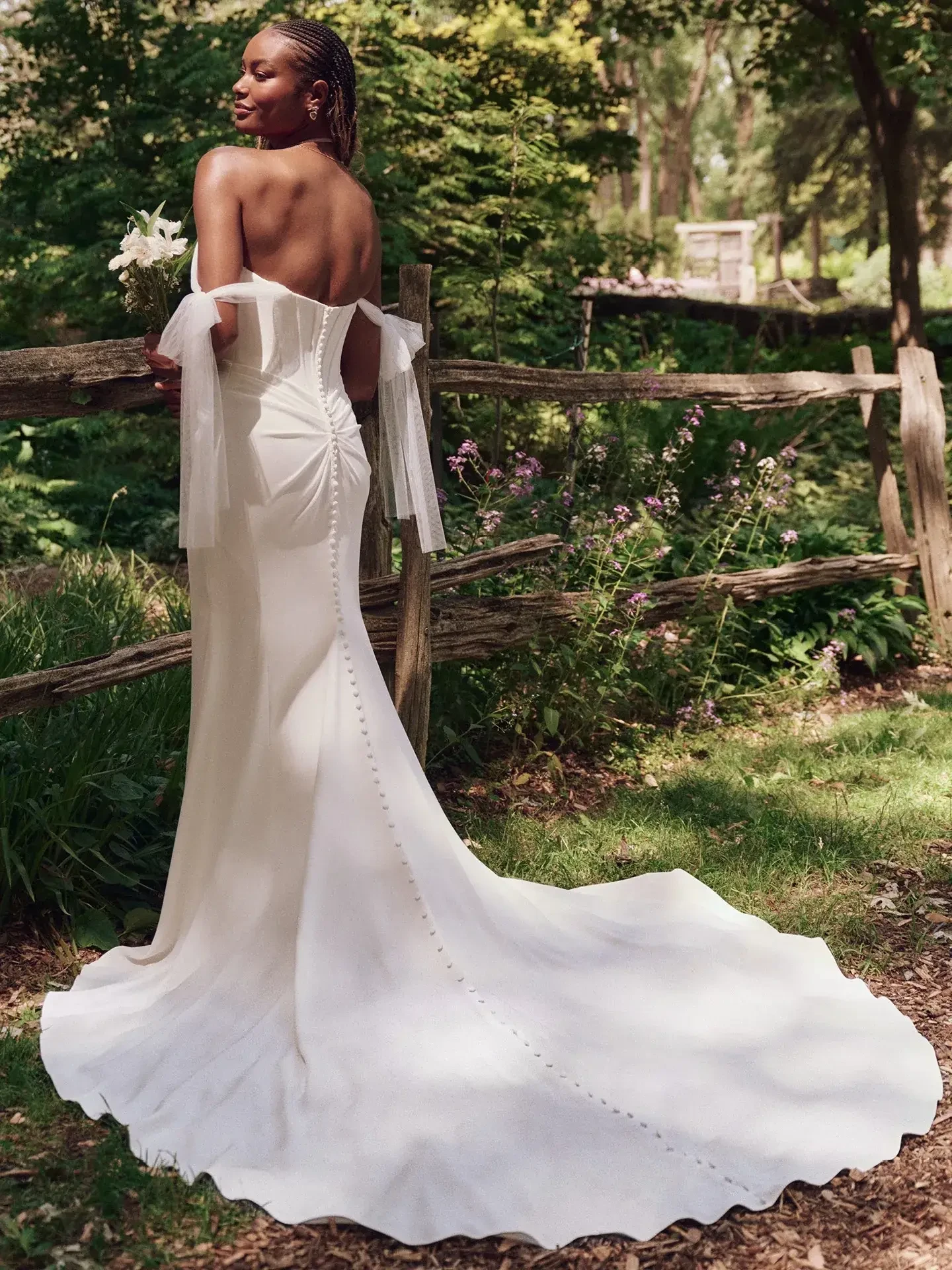 A woman in a white off-shoulder wedding gown stands elegantly in a lush garden, holding white flowers, exuding joy and serenity.