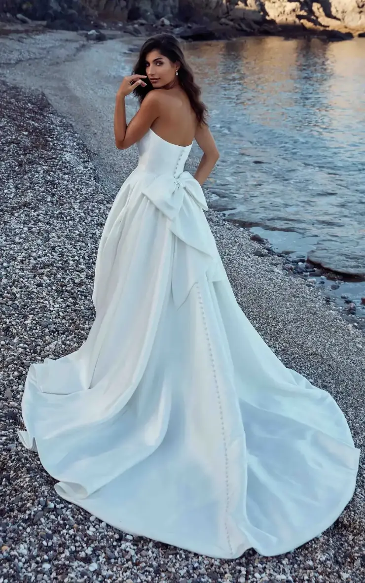 Model is wearing a pearl ivory Mikado ballgown with a cat-eye neckline, basque waist, attached back bow, and pockets.