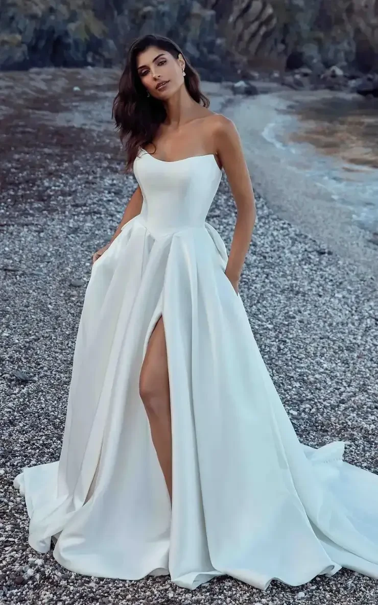 Model is wearing a pearl ivory Mikado ballgown with a cat-eye neckline, basque waist, attached back bow, and pockets.