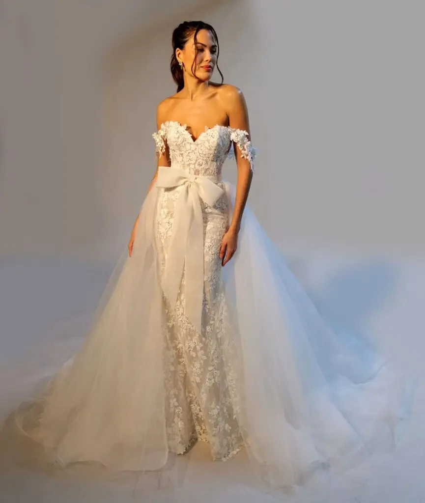 A woman in an elegant, off-shoulder lace wedding gown with a large bow and tulle skirt. She looks serene, standing against a simple background.