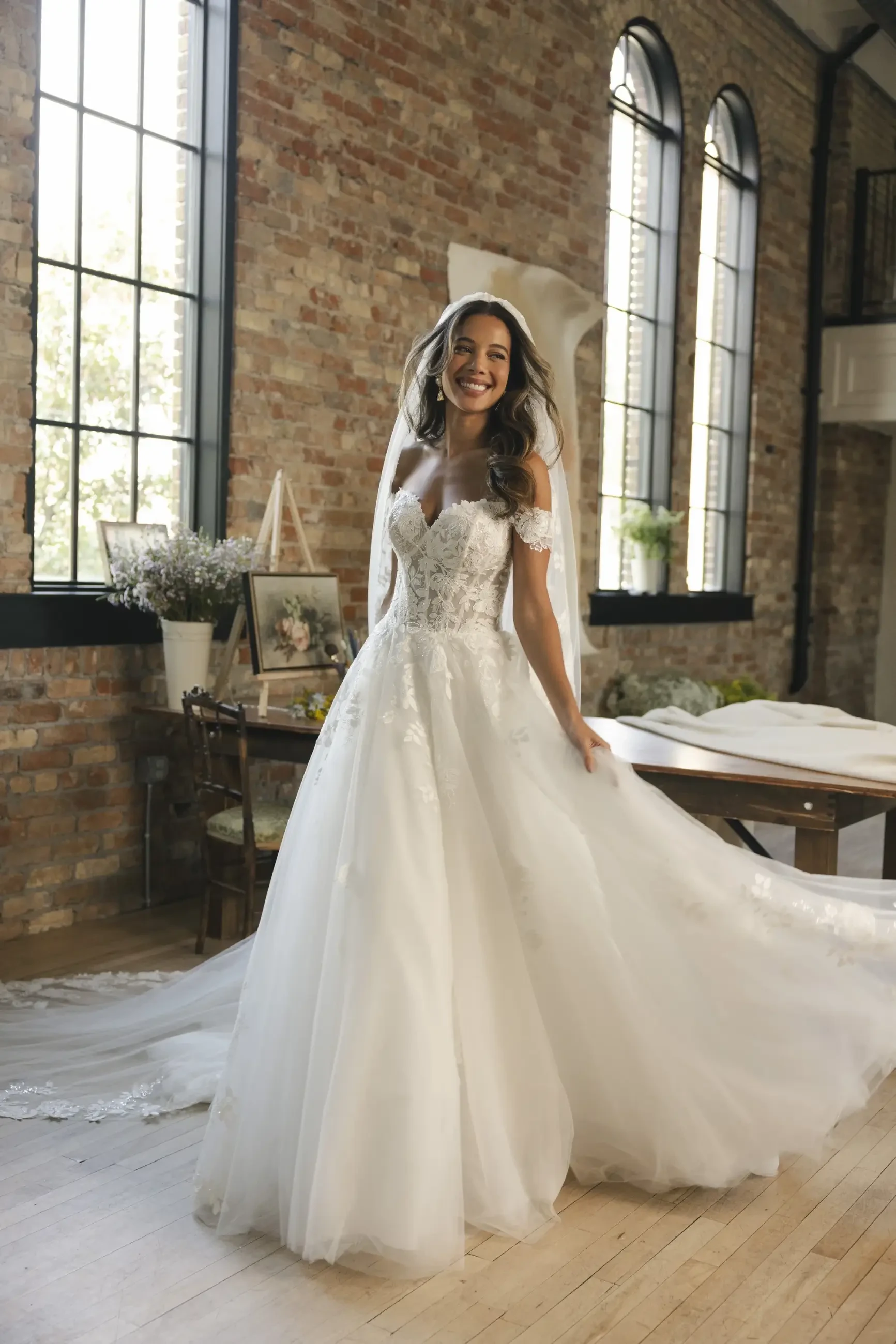 Bride in lace wedding dress stands in sunlit room with rustic brick walls and large windows, smiling joyfully. Elegant and serene atmosphere.