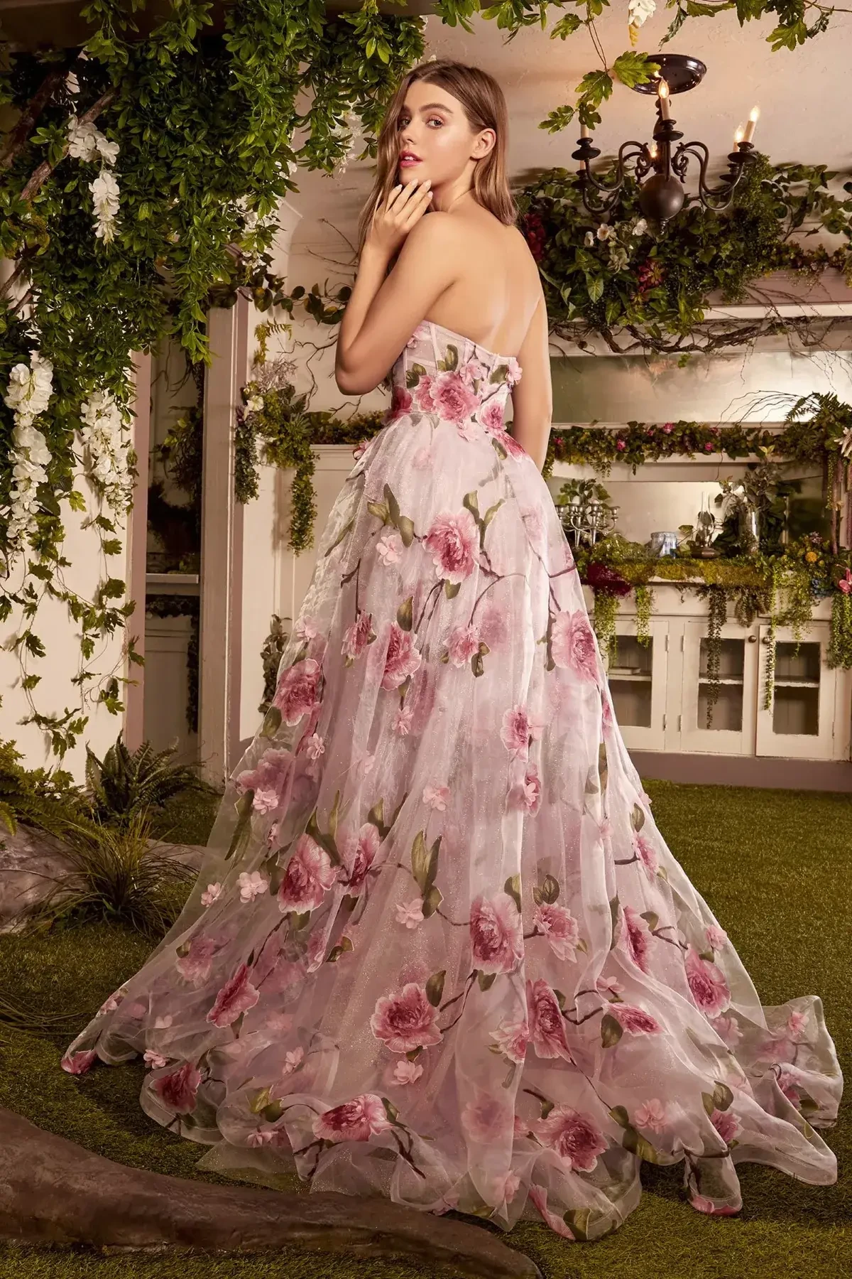 Model is wearing a strapless A-line organza gown with a sheer corset bodice, pink rose print, dimensional floral appliqués, and a front leg slit.