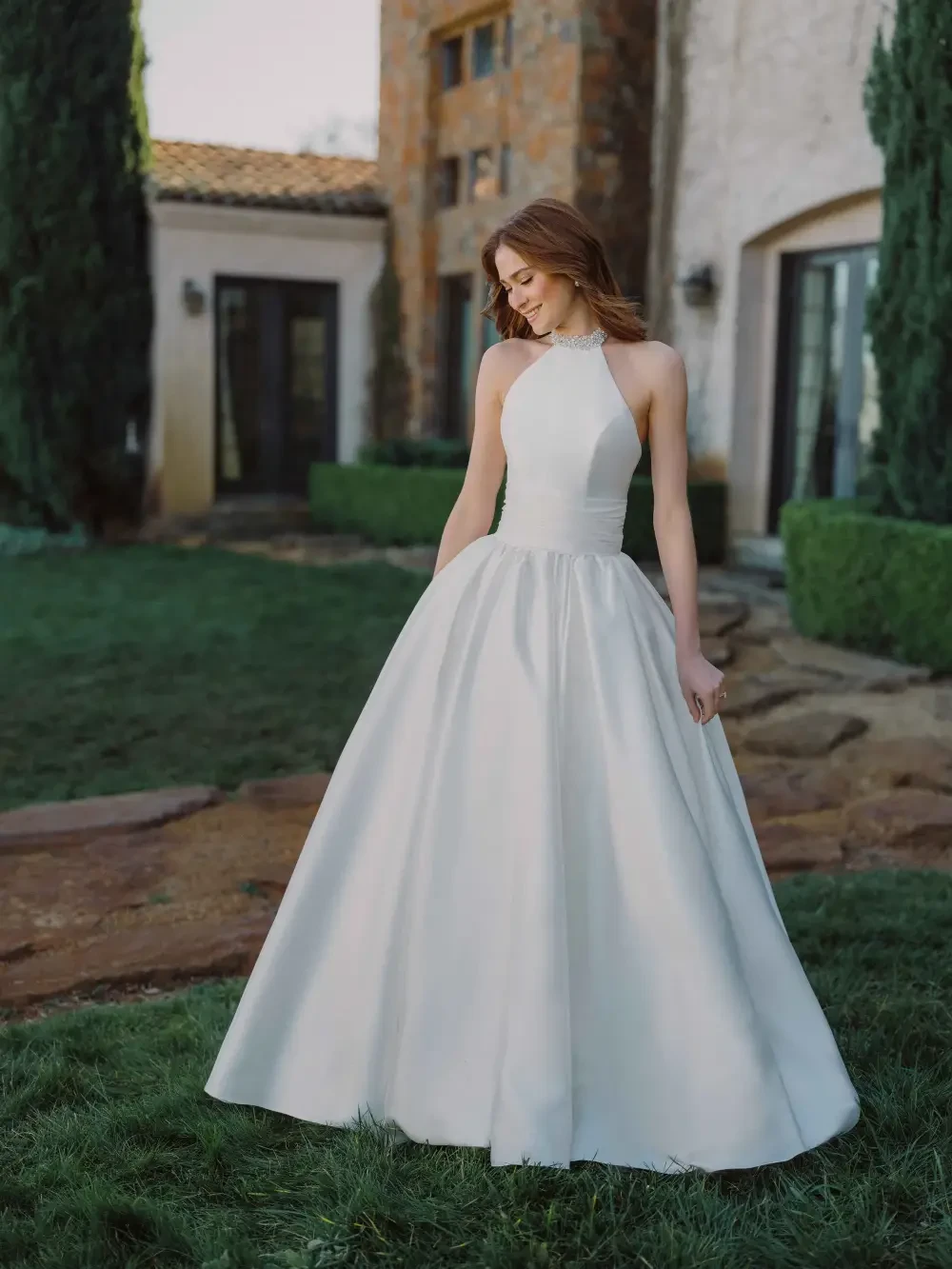 A woman in a halter-neck white gown standing on green grass in front of a stone building with trees in the background.