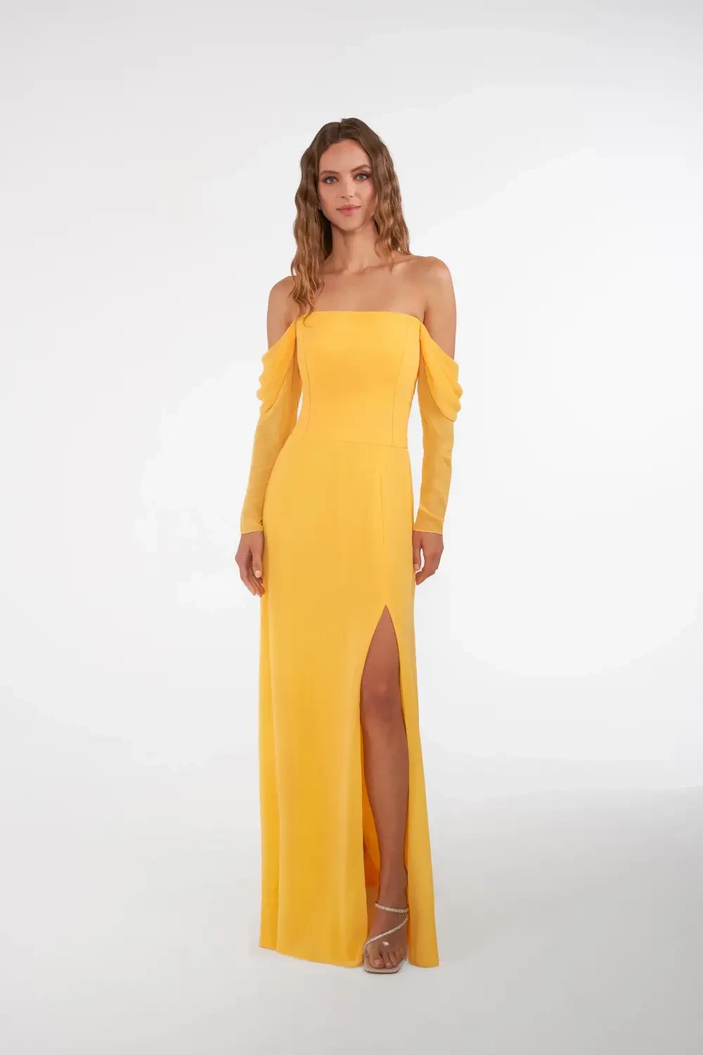 A woman in a long yellow off-the-shoulder dress with a thigh-high slit and long sleeves, standing against a plain background.