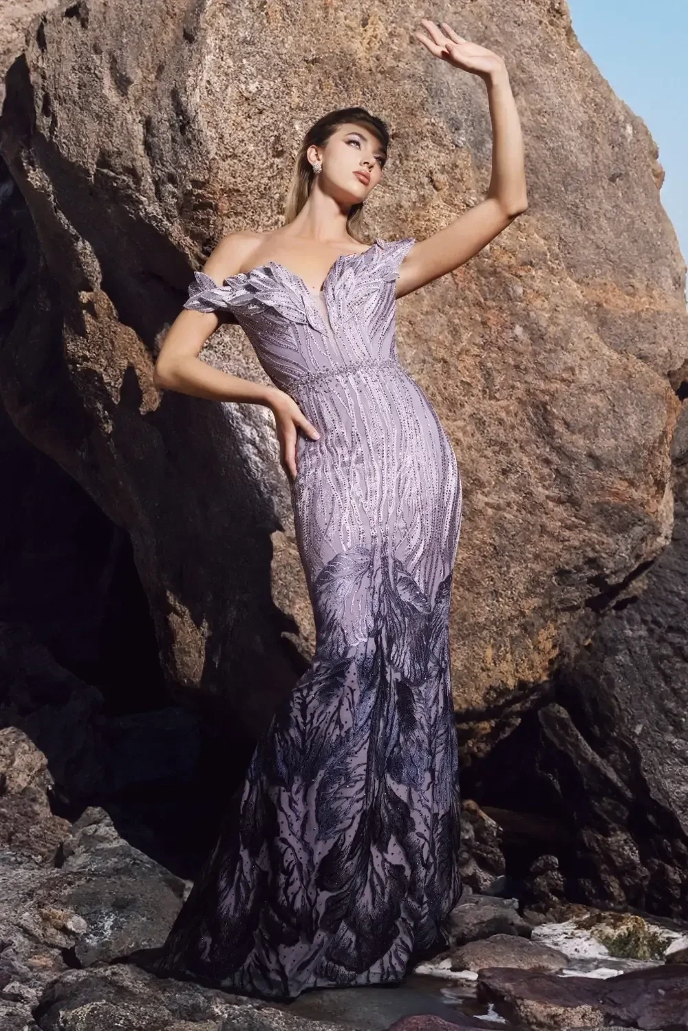 A model wearing a luxurious off-the-shoulder gown that transitions from light gray to black, featuring intricate details and a fitted silhouette, standing against a backdrop of lush green foliage.