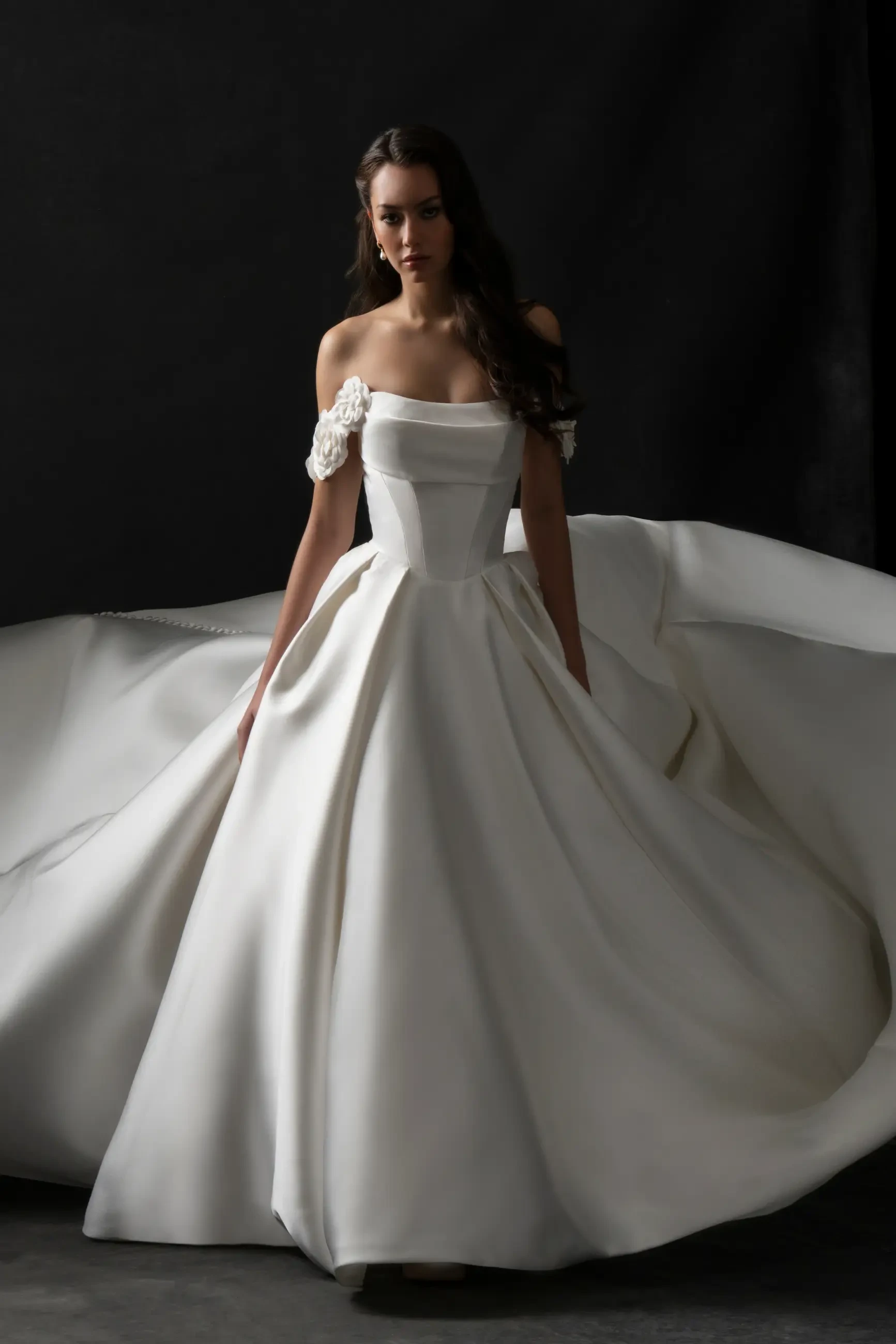 A model wearing a white off-the-shoulder gown with a flowing skirt, posed against a dark background.