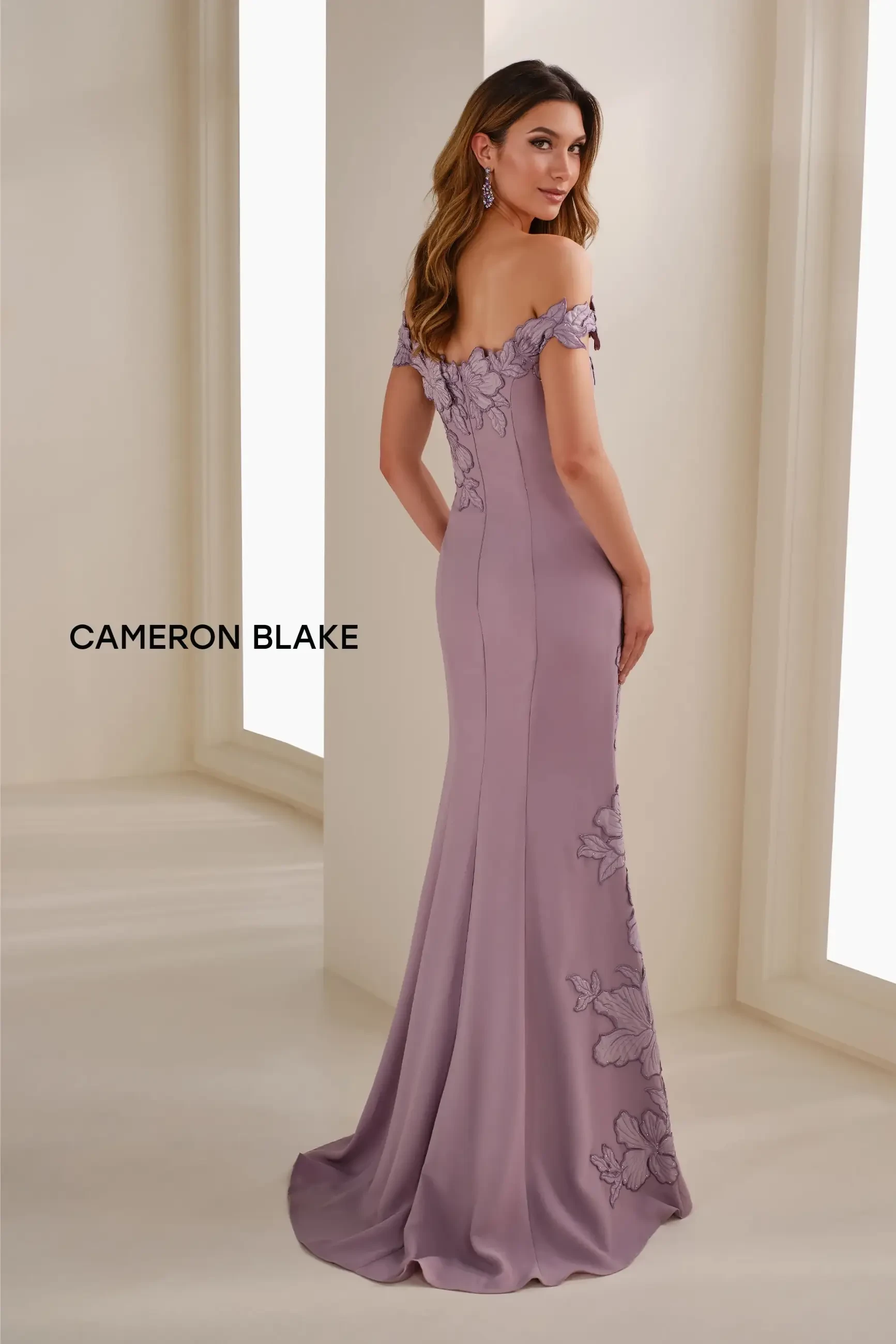 Model is wearing the Cameron Blake CB901, a heather purple curved-neck off-the-shoulder mermaid dress with embroidered flower appliqué, subtle stone accents, and a coordinating crepe shawl.