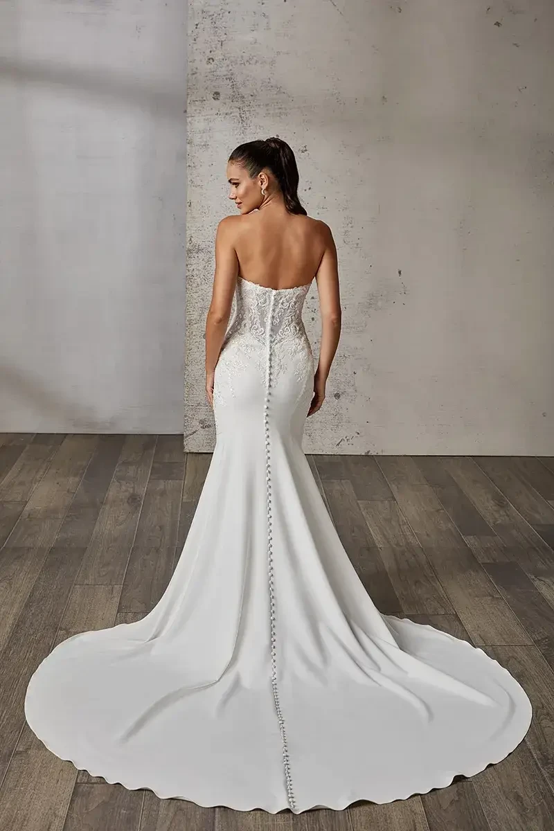 A woman in a strapless white gown with a fitted silhouette and a train, viewed from the back. The gown features intricate detailing on the bodice and a row of buttons running down the back.