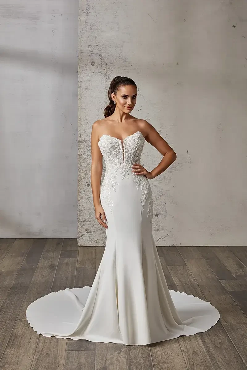 A woman in a strapless wedding dress with intricate beadwork, standing against a minimalist background.
