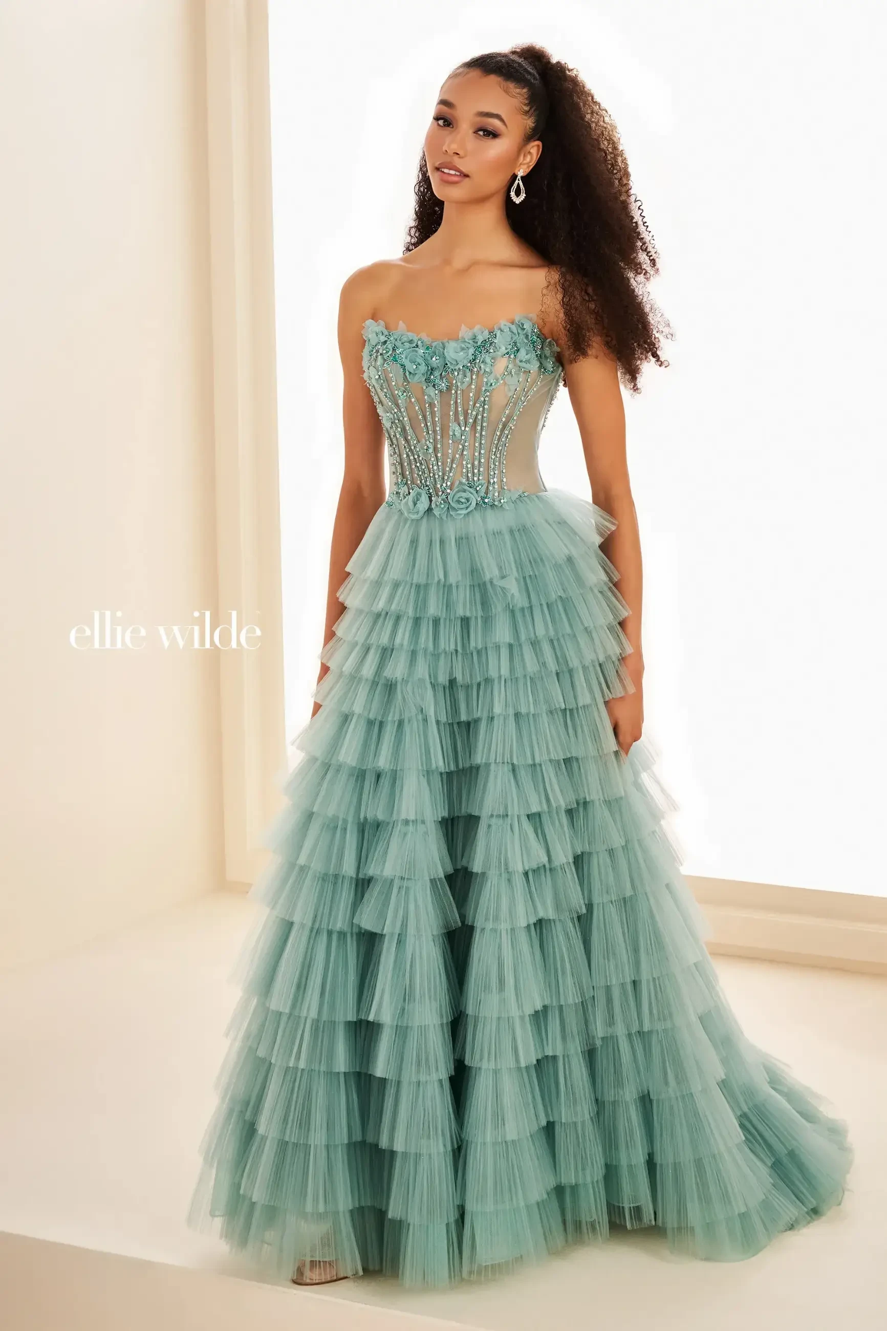 A woman wearing an elegant seafoam green gown with layered tulle, floral detailing, and a fitted bodice stands gracefully, exuding sophistication.