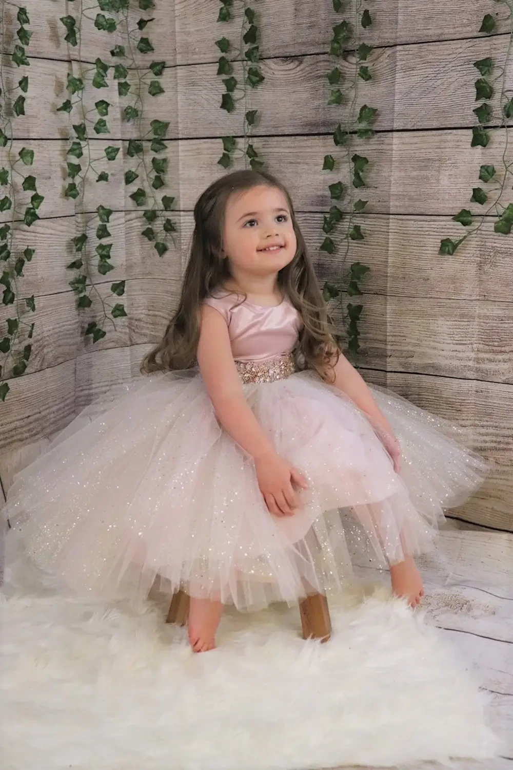 A young girl in a sparkly pink dress sitting on a wooden chair in front of a wooden wall with greenery.