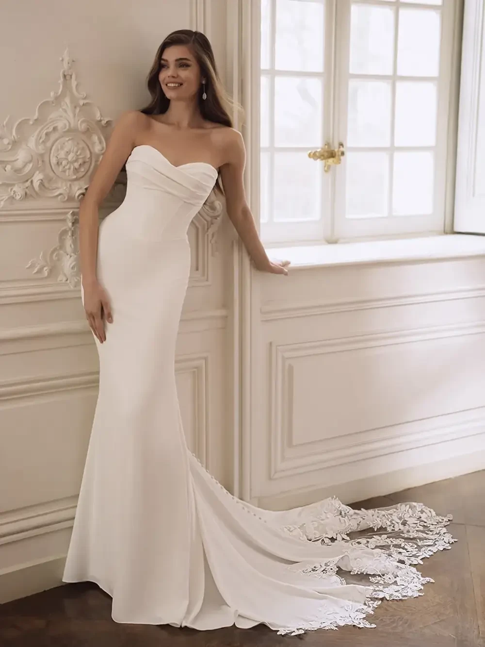 A woman in a strapless white wedding dress stands near a window, showcasing a fitted silhouette with a lace train.