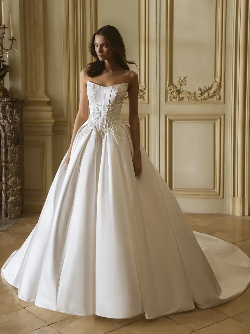 A woman in an elegant, strapless white wedding gown with intricate embroidery stands in a luxurious room with ornate walls, exuding grace and sophistication.