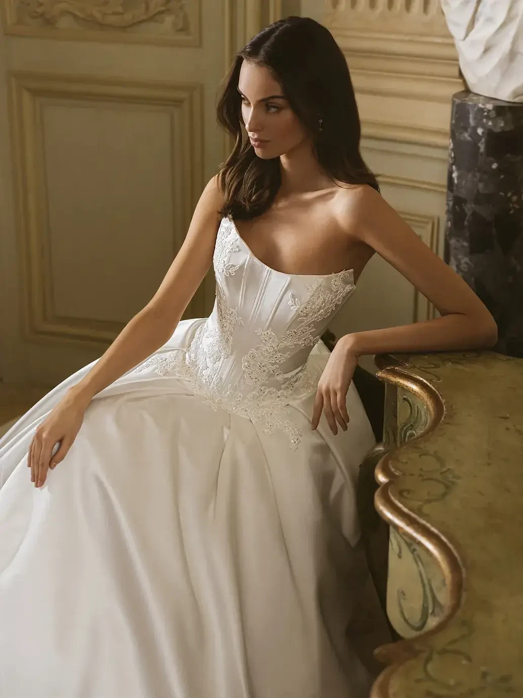 A woman in a strapless, white wedding dress elegantly sits beside an ornate table in a luxurious setting.