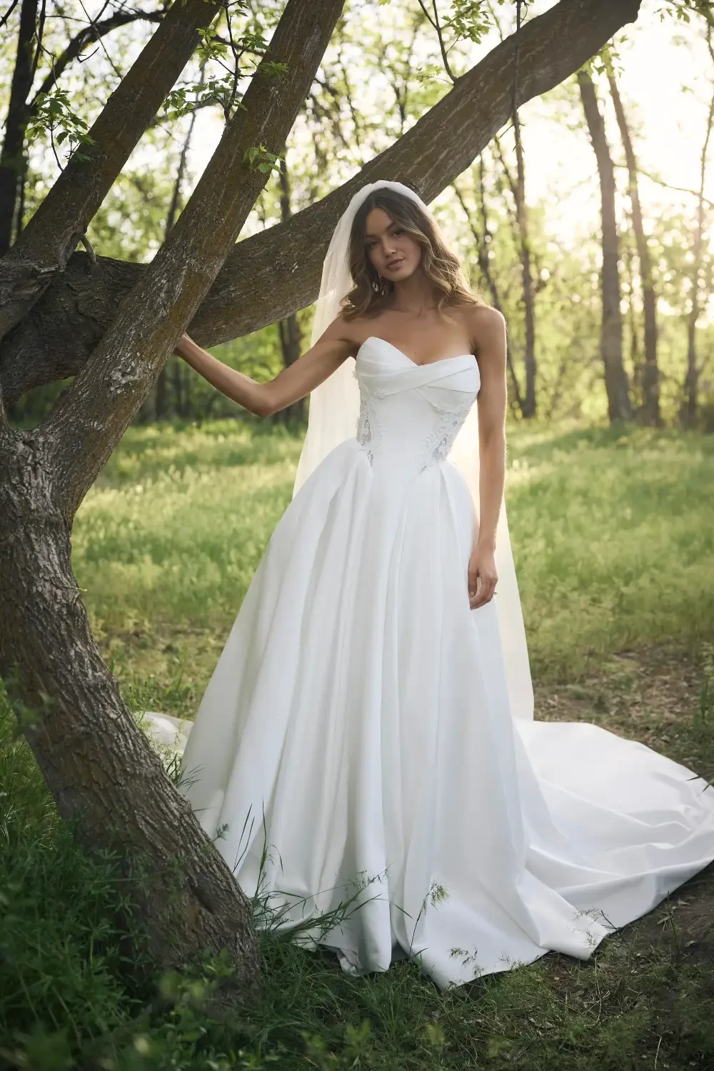 A woman in a white wedding dress standing under a tree in a green, sunny setting.