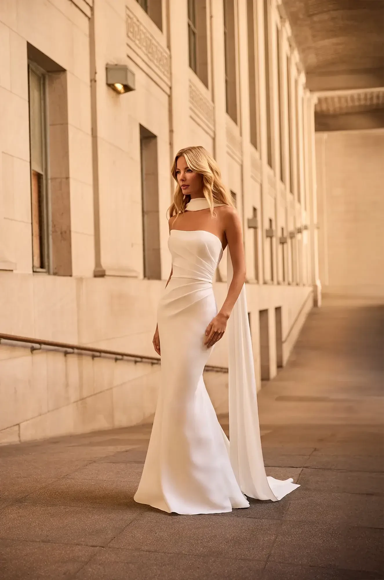A woman in a sleek, strapless white gown with a long train stands confidently in an elegant, sunlit corridor lined with columns. The setting feels timeless and sophisticated.