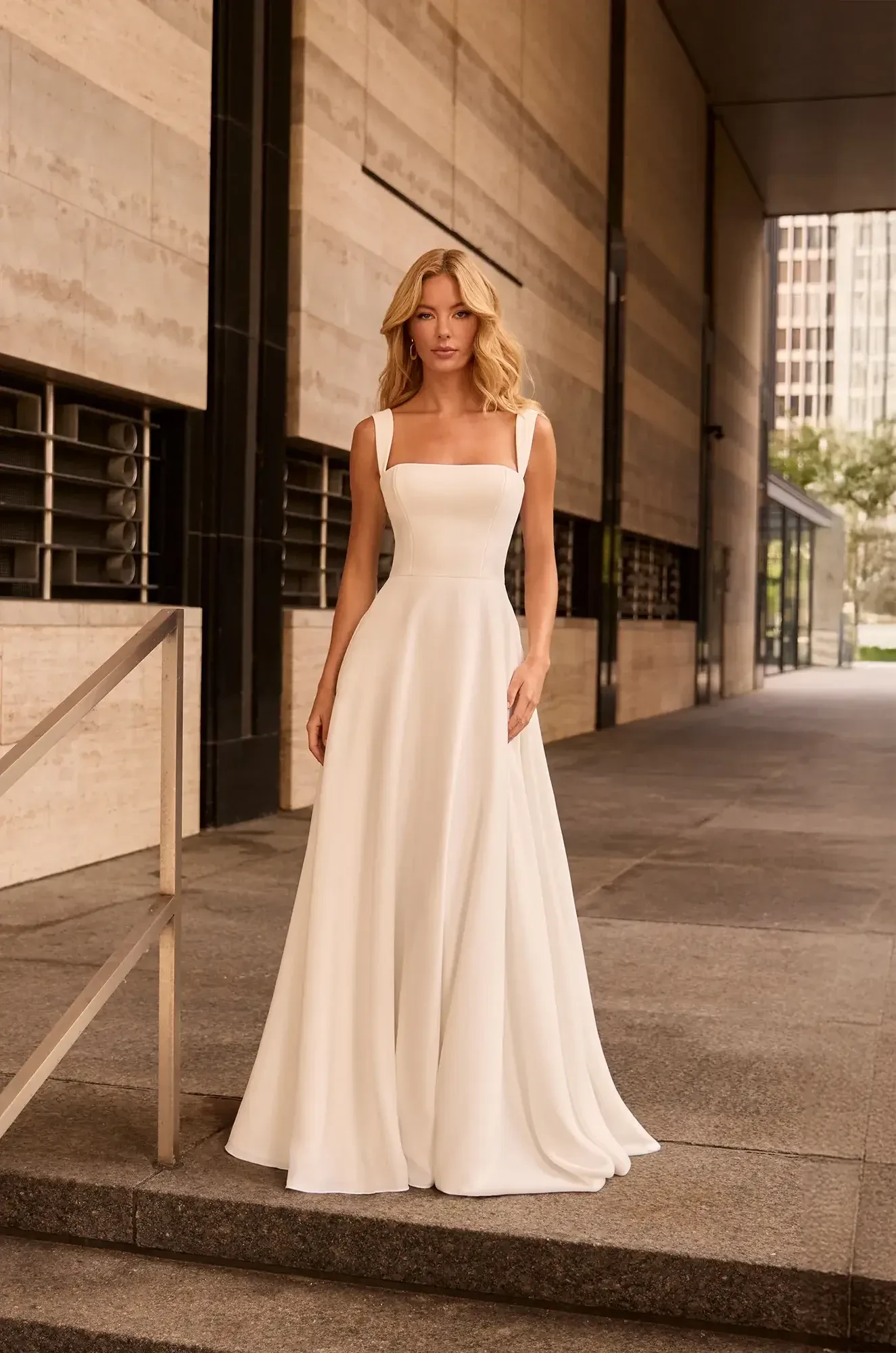 A woman in an elegant white gown stands confidently in an urban setting with modern architecture. The mood is sophisticated and serene.