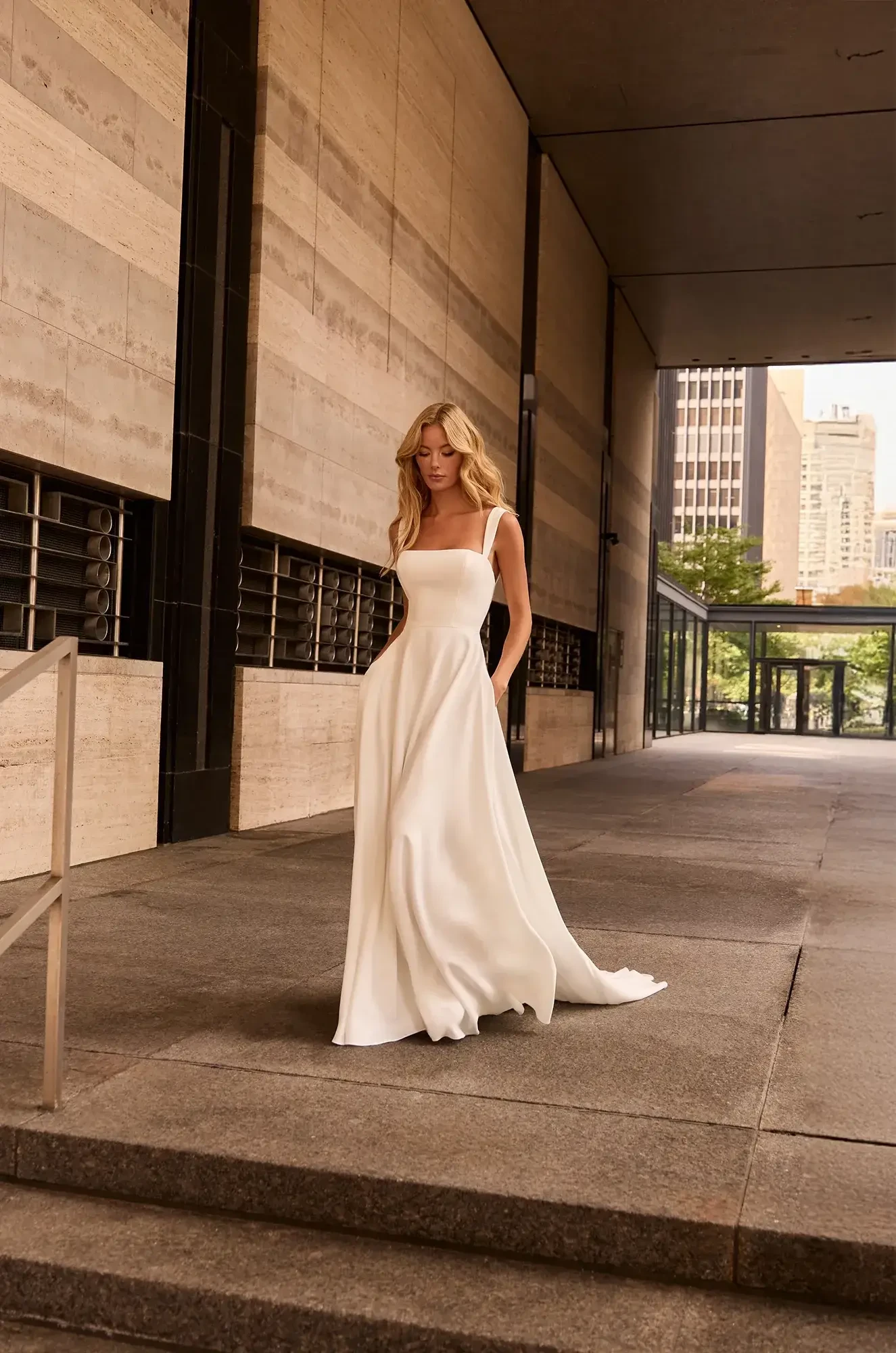 A woman in a white, elegant, flowy dress walks through a modern architectural walkway. The scene is urban and serene, with sunlight filtering through.