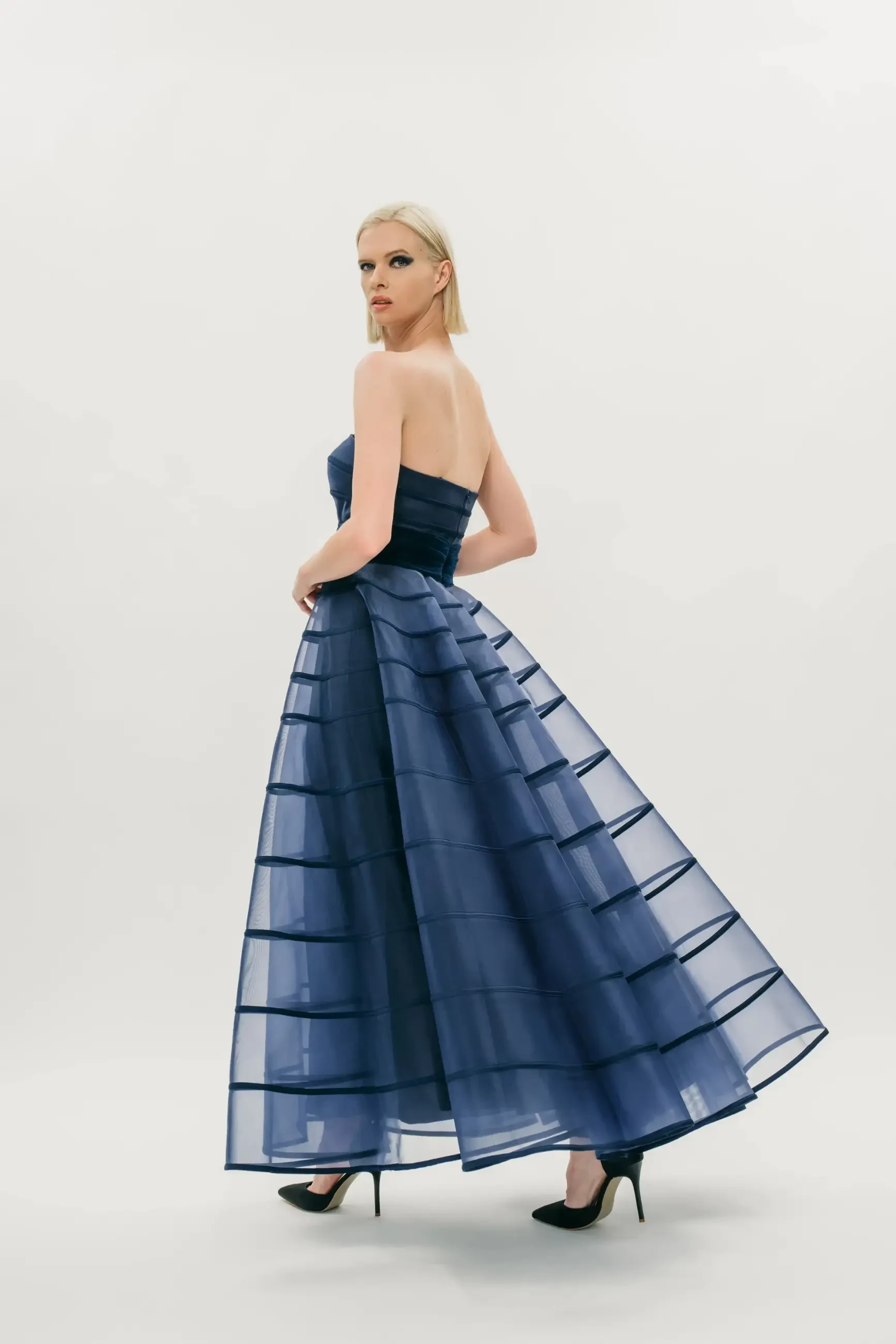 Model is wearing a strapless A-line gown with a smooth structured bodice, cummerbund waist, and softly pleated flowing skirt.