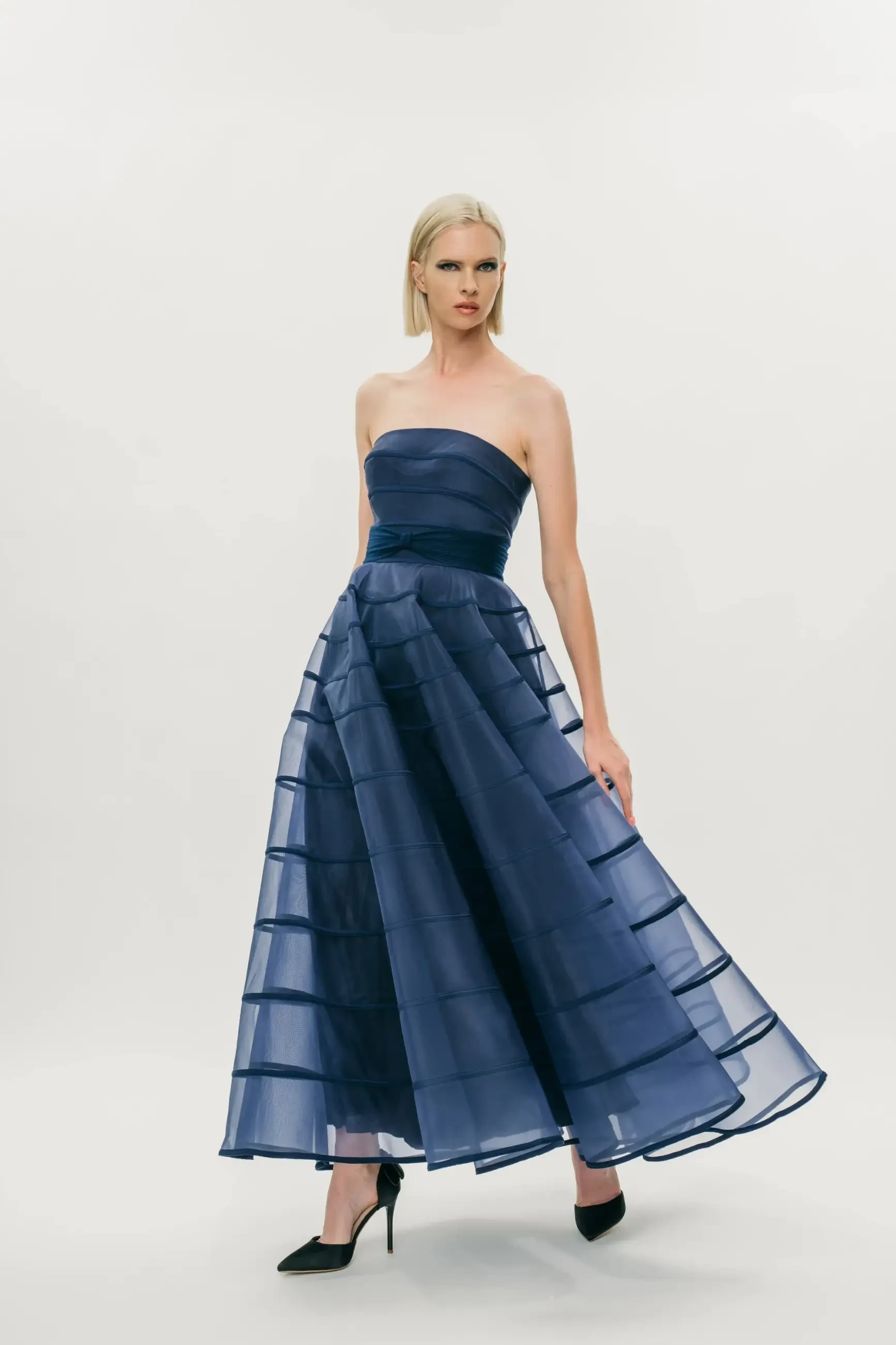 Model is wearing a strapless A-line gown with a smooth structured bodice, cummerbund waist, and softly pleated flowing skirt.
