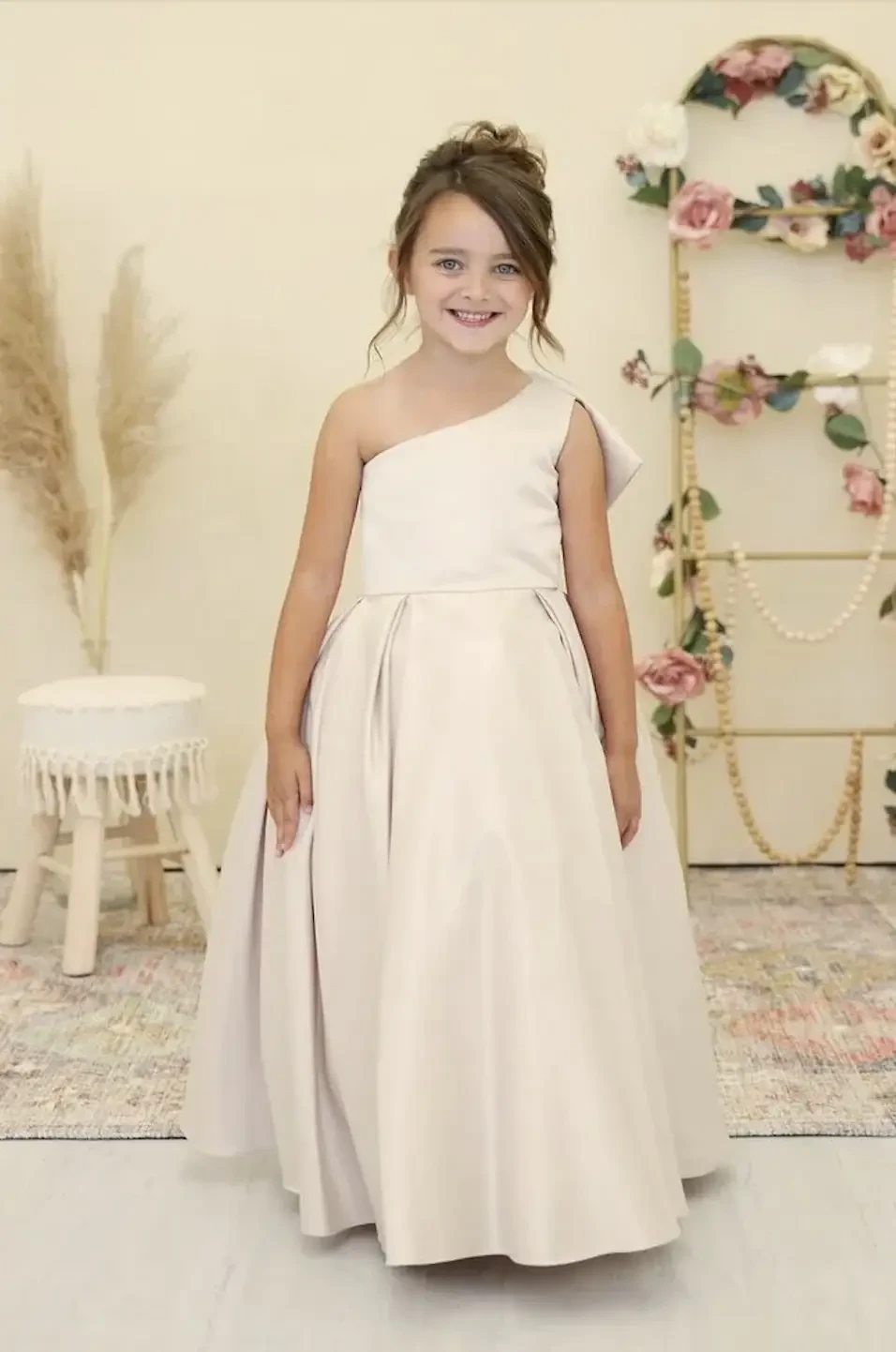 A young girl wearing a one-shoulder, knee-length cream dress, smiling in a decorated indoor space.