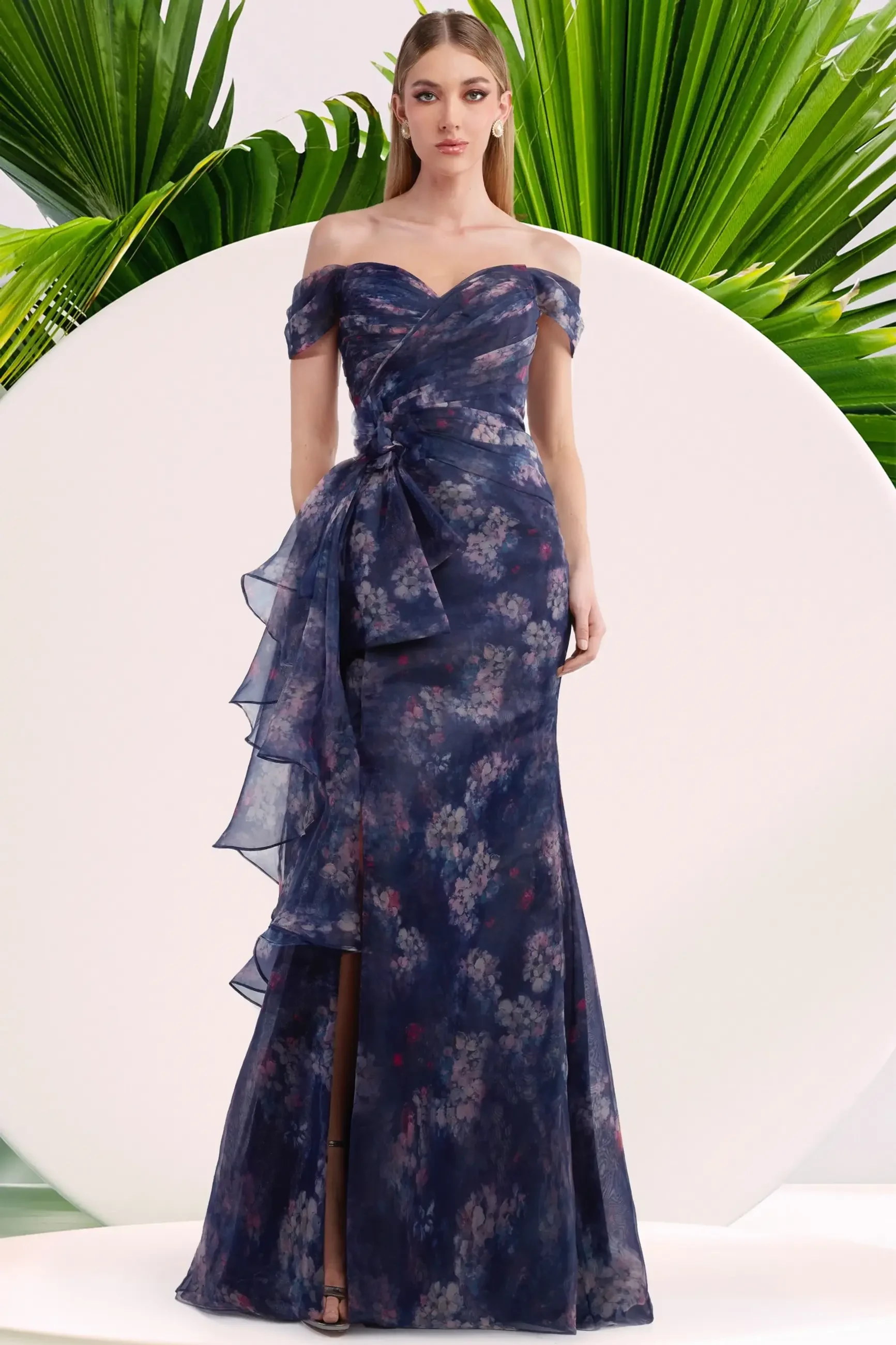 A woman in an off-the-shoulder floral dress with a mermaid silhouette and a side slit, standing against a backdrop of green tropical leaves.